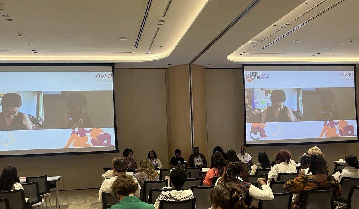 A picture of a panel discussion at AWID International Forum 2024,  in a modern meeting room. Four speakers are seated at a table, with one moderator seated beside them, and another moderator joining them through Zoom, visible on a large screen behind them. The audience, mostly women, is seated in rows, facing the panel. The room is well-lit, with a contemporary design.