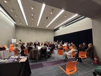 A roomful of diverse participants seated in different round tables, with everyone paying attention to a speaker at the front of the room.